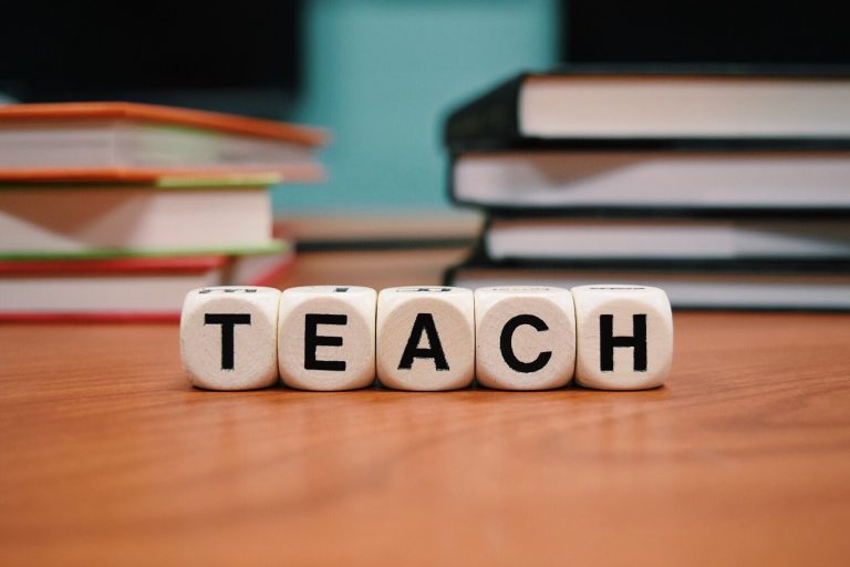 Close-up of lettered blocks spelling "TEACH" on a wooden surface, with stacks of books blurred in the background, evoking the supportive atmosphere of an independent school.