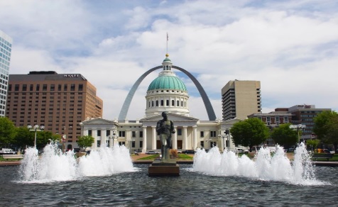 A large fountain with multiple jets in front of a statue, the Old Courthouse with its green dome, and the iconic Gateway Arch in Missouri stand beneath a partly cloudy sky in St. Louis.