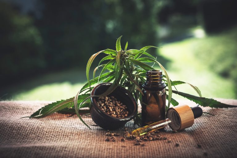 A bottle of CBD oil with a dropper, a bowl of seeds, and a cannabis plant in the background are arranged on hessian cloth, hinting at natural or herbal remedies, possibly CBD or hemp products.