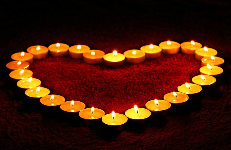 A group of small tealight candles arranged in the shape of a heart, glowing warmly on a textured red surface in a dark setting, creates a touching tribute to mark the death of a pet.