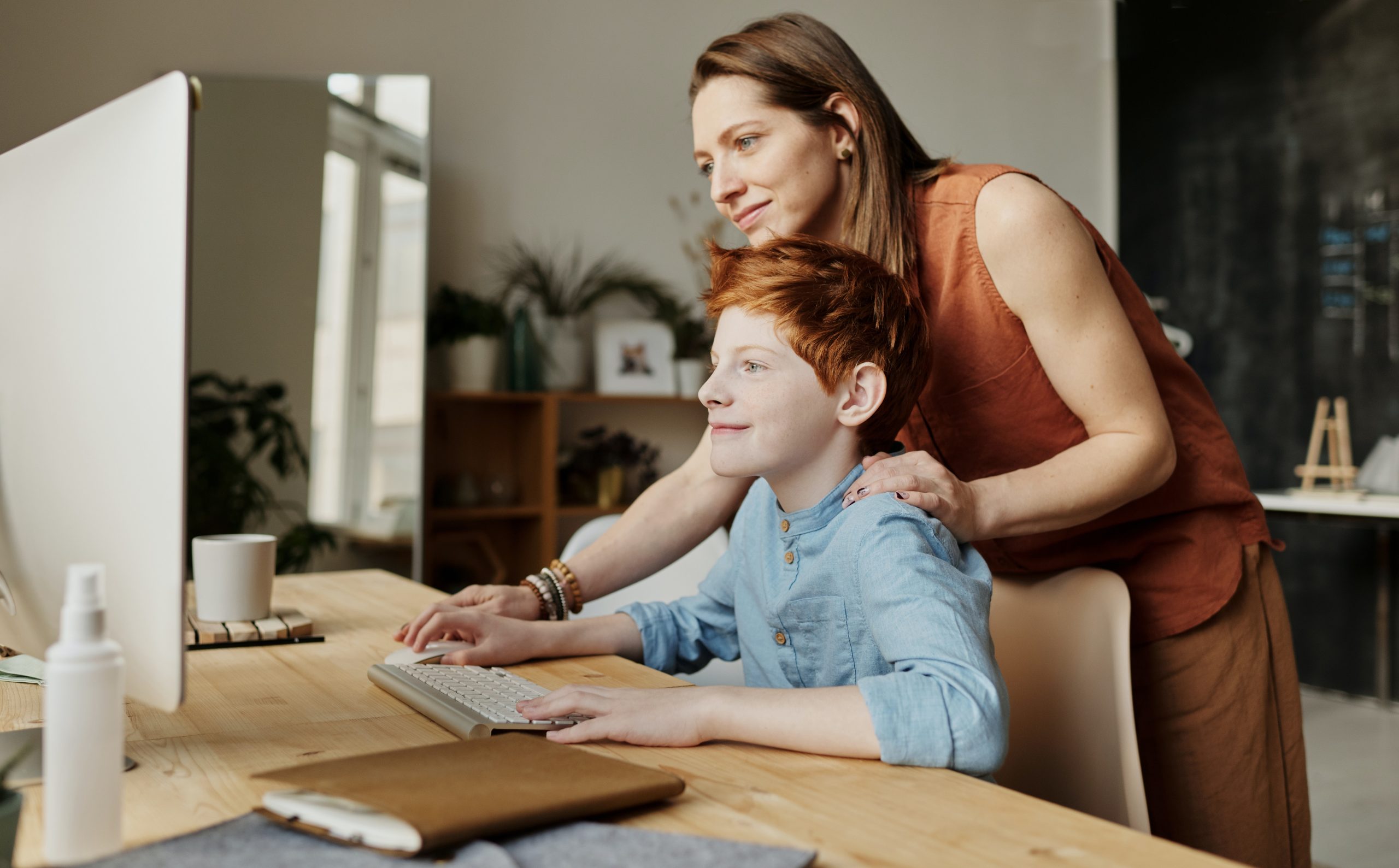 A boy sat at a computer with a woman stood next to him with a hand on his shoulder. She is also loking at the computer screen