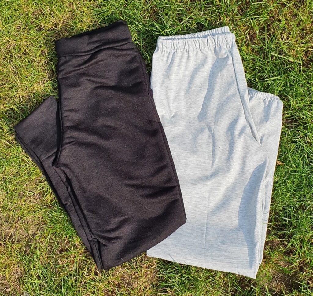 Two pairs of tracksuit bottoms, one black and one light grey, are laid out side by side on green grass in bright sunlight—perfect wardrobe must-haves for comfort and style.
