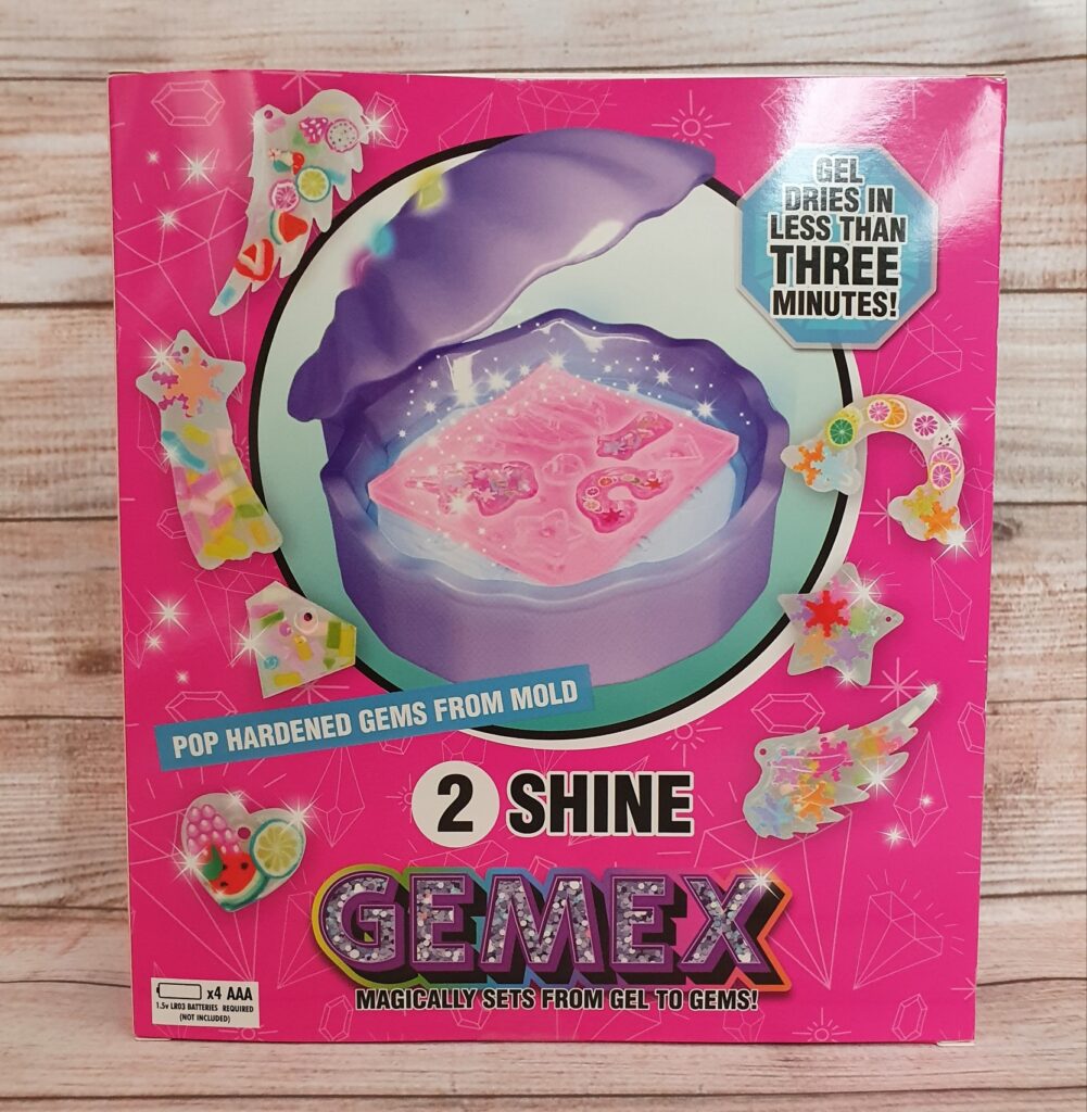 A pink box labelled "GEMEX 2 Shine" displays a mould with pink gems, colourful fashion toys gem designs, and text saying "Gel dries in less than three minutes!" and "Pop hardened gems from mould." The background features a wooden surface.