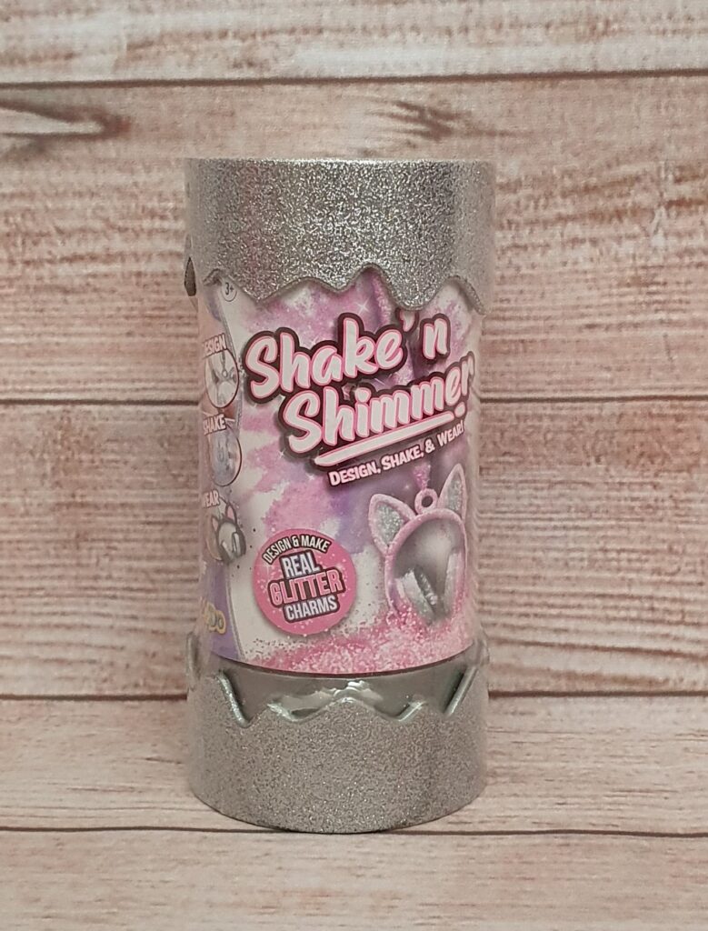 A silver cylindrical container labelled "Shake 'n Shimmer" with pink and white text, featuring sparkly designs and images of glitter charms, highlights trendy fashion toys against a wooden background.