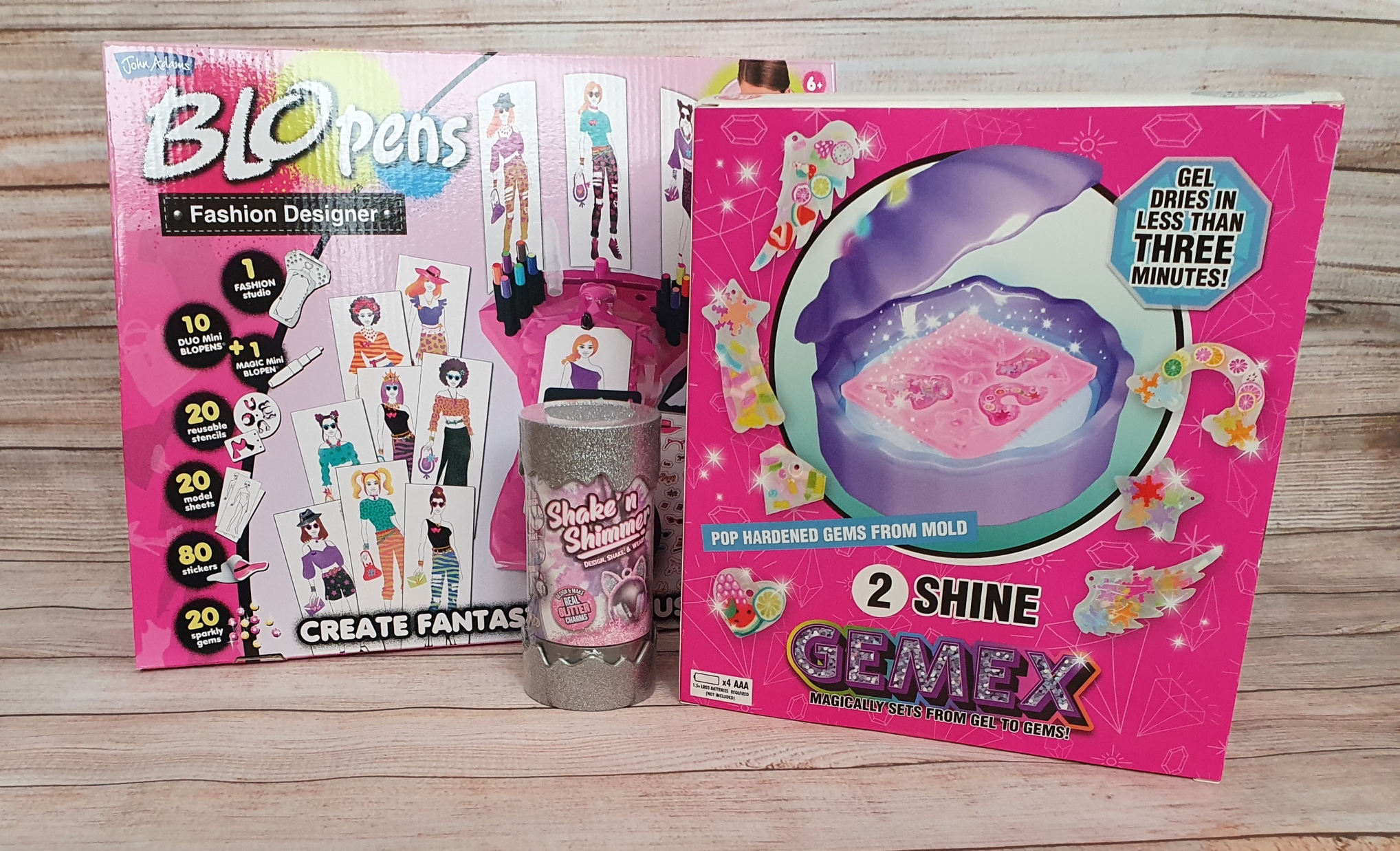 Two colourful fashion toys in pink packaging are displayed: one is a BLOpens Fashion Designer kit, and the other is a Gemex 2 Shine gem-making set with a shaker bottle in front of them, all on a wooden background.