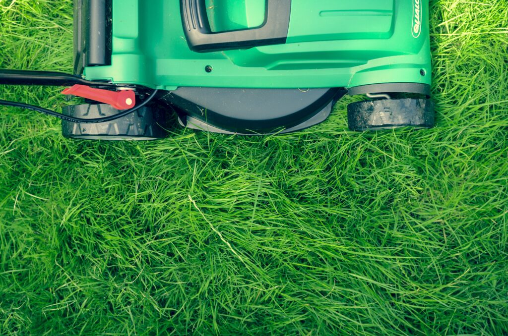 A close-up, top view of a green lawnmower in use on tall, green grass, showing the mower partially cutting the grass with a visible handle and wheels—perfect for caring for your garden.