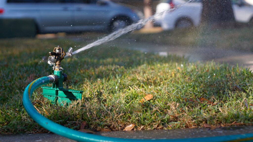 A green garden sprinkler attached to a hose sprays water over a patch of grass, showing the importance of caring for your lawn, with parked cars blurred in the background.