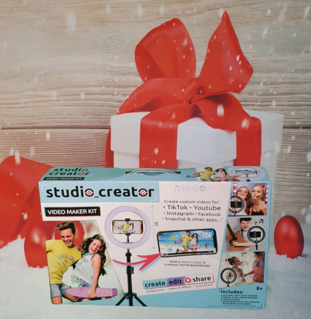 A Studio Creator video maker kit box is displayed in front of a large white gift box with a red ribbon, set against a wooden background with red baubles—one of the must-have gifts for children Christmas 2020.