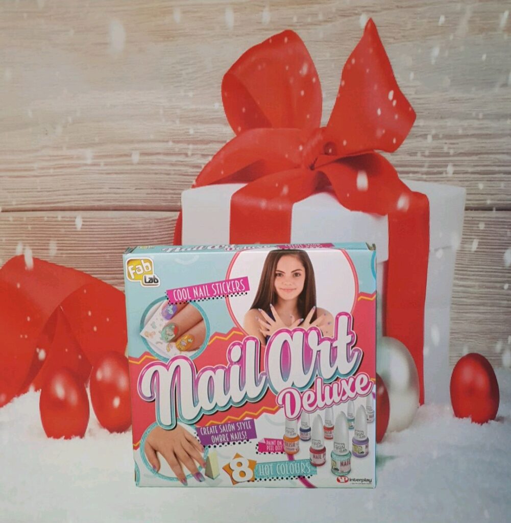 A Fab Lab Nail Art Deluxe kit box is displayed in front of Christmas presents with red bows and baubles. Snowflakes fall, creating a festive scene—one of the must-have gifts for children Christmas 2020.