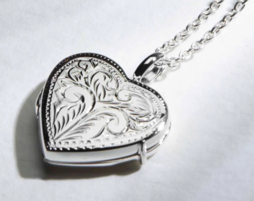 A silver heart-shaped locket with intricate floral engravings hangs from a delicate chain, making it one of the sweetest Mother’s Day gift ideas 2021, shown resting on a white surface.
