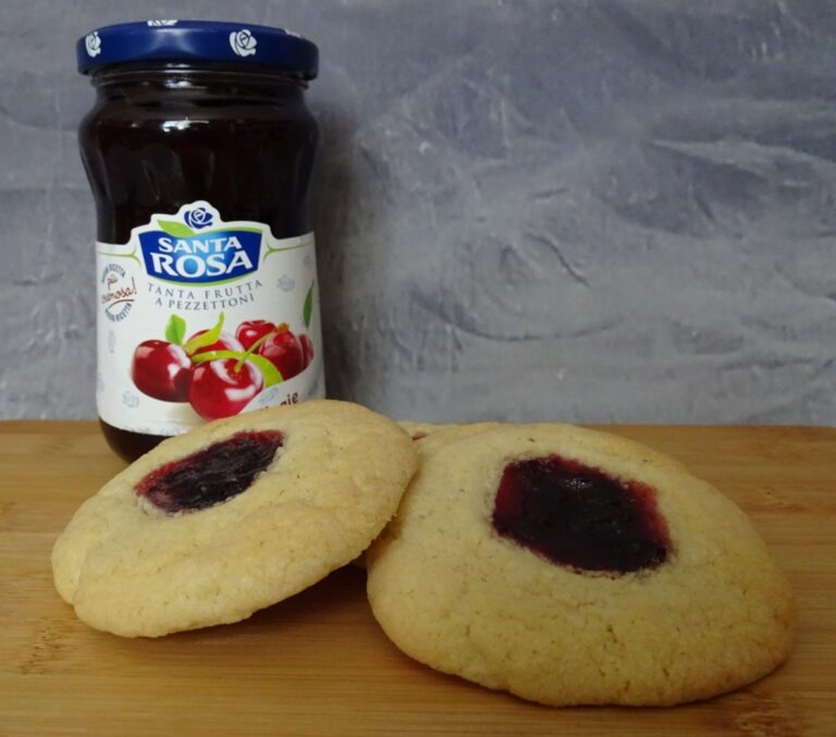 Two thumbprint biscuits with red jam centres are placed on a wooden surface in front of a jar of Santa Rosa cherry jam against a grey background, perfect for trying out your favourite Cherry Thumbprint recipe.