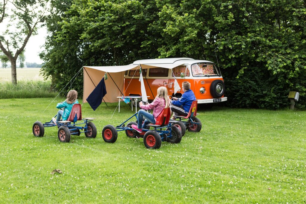 Children playing on pedal go karts next to an orange VW campervan with an awning