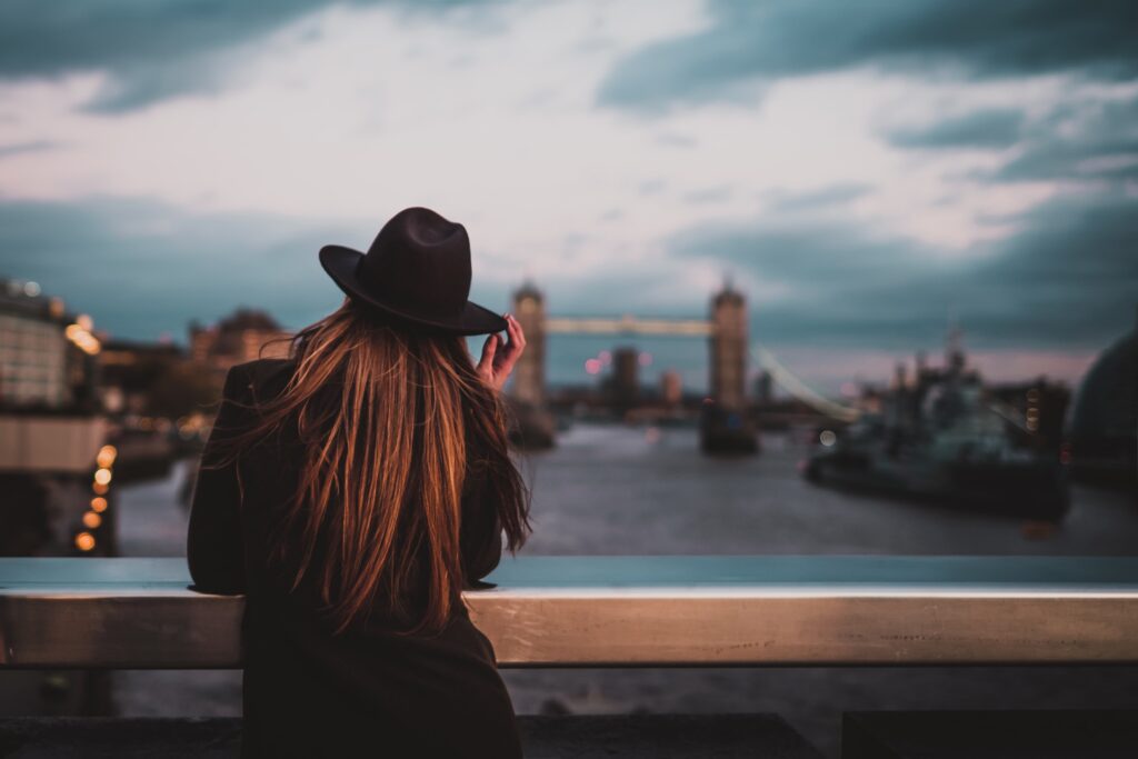 A person with long hair wearing a black hat and coat leans on a railing, gazing at the Thames and Tower Bridge in London at dusk—an ideal scene that hints at why a serviced apartment is perfect for entertaining guests in style.