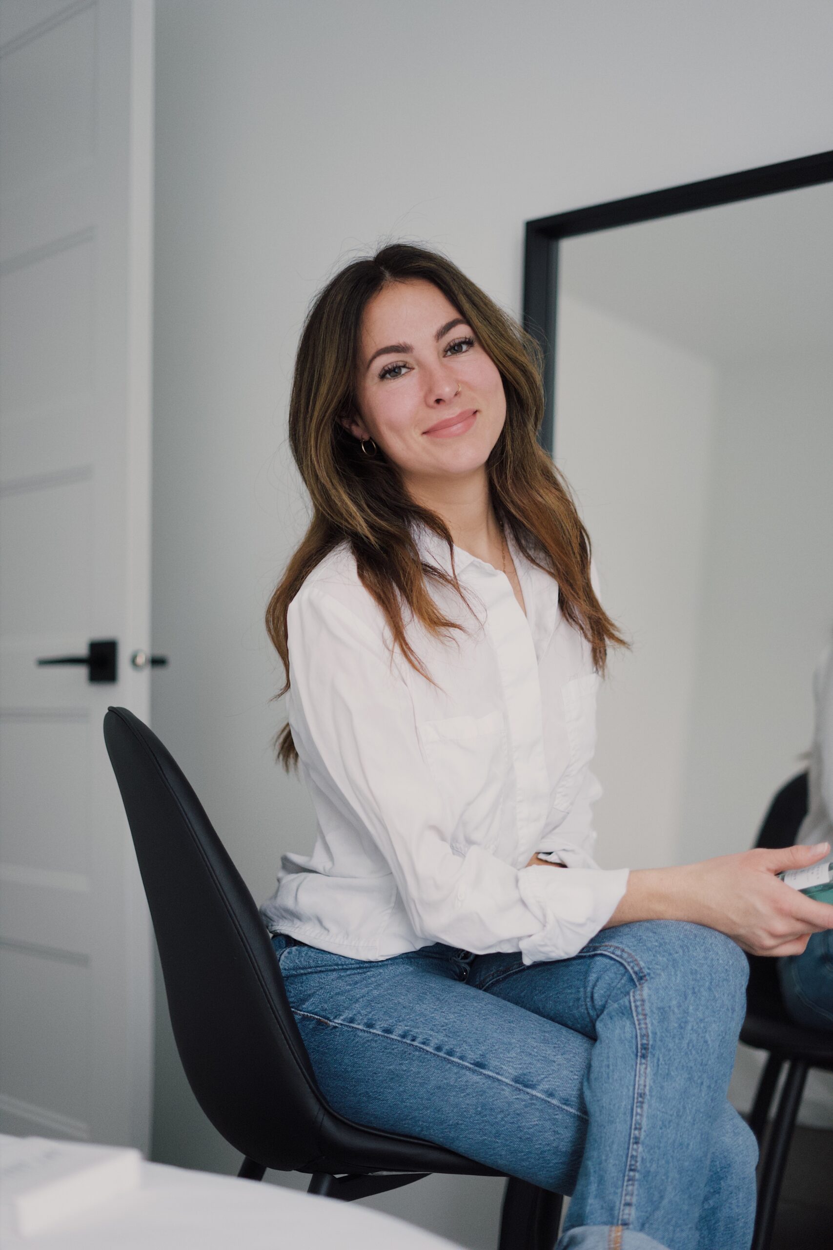A woman with long brown hair, wearing a white shirt and blue jeans, sits smiling in a modern, minimalistic room—showcasing why a serviced flat is perfect for entertaining guests with its stylish mirrors and bright white walls.