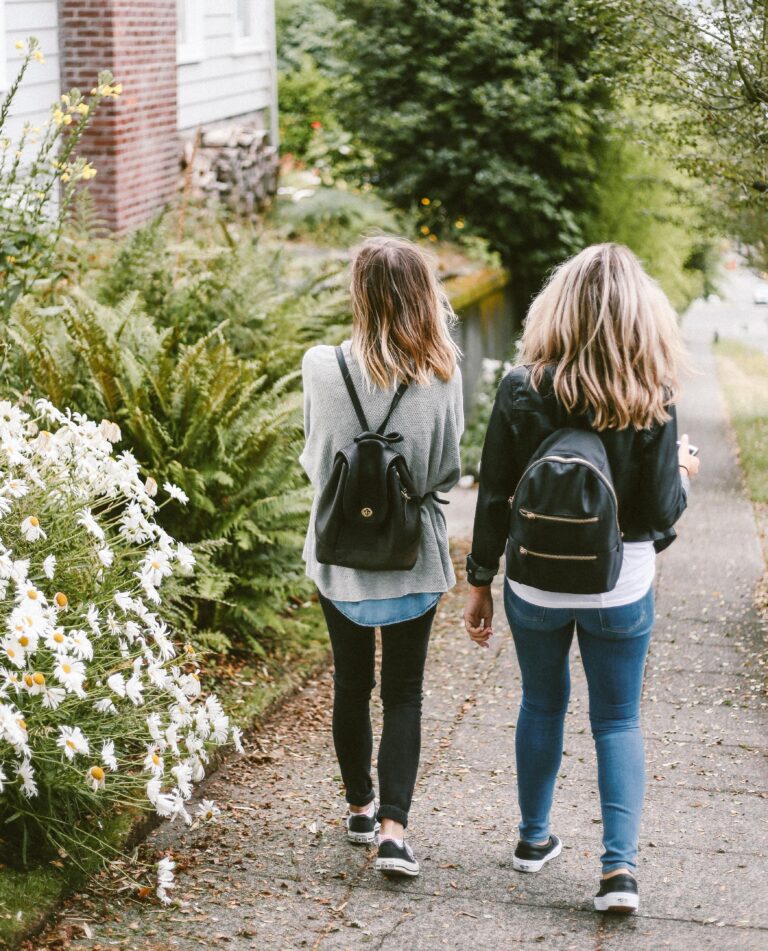 Two people with rucksacks walk side by side down a pavement lined with green plants and white flowers—capturing the spirit of adventure and The Benefits of School Trips for Children. Both have shoulder-length hair and are dressed casually.