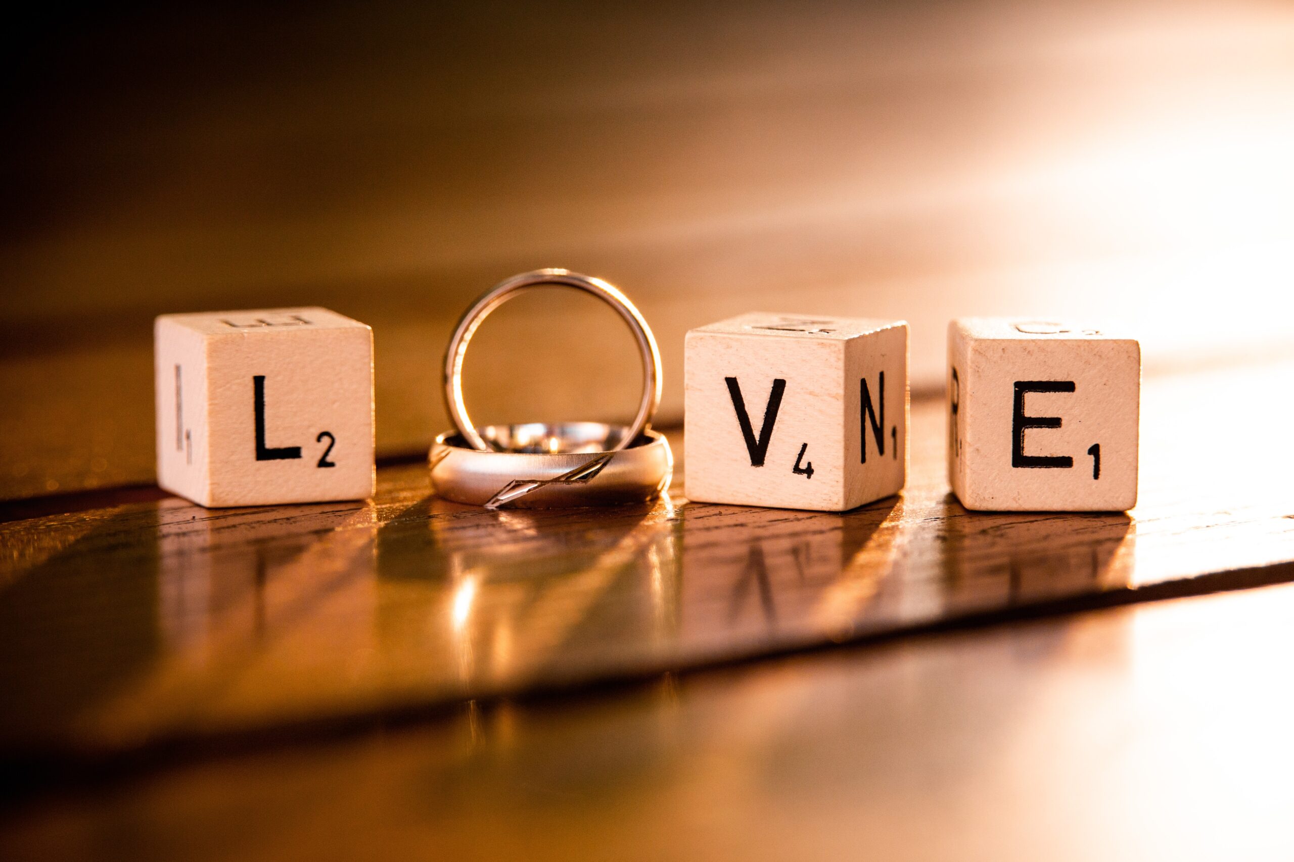 Four Scrabble tiles spelling "LOVE" are arranged in a line, with wedding rings standing upright between the tiles, all set on a wooden surface with warm lighting.