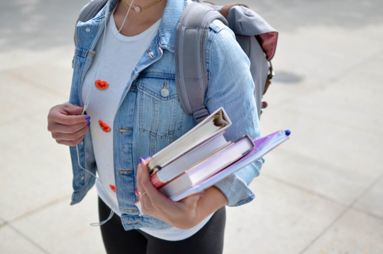A person wearing a denim jacket and rucksack holds three books, including A Parents' Guide to A Levels, and some papers while walking outdoors. Earbuds are visible, with only the torso shown.