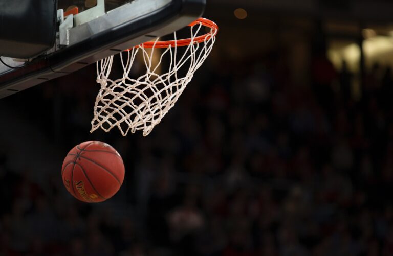 A basketball is about to pass through a hoop, captured mid-air during a Top NBA 2022 moment, with a blurred background of spectators in an indoor arena.