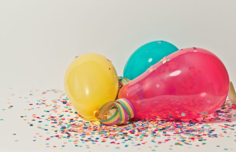 Three colourful balloons—yellow, turquoise, and pink—lie on a white surface scattered with multicoloured confetti, creating a festive vibe that’s perfect inspiration for how to make a healthy party menu fun and inviting.