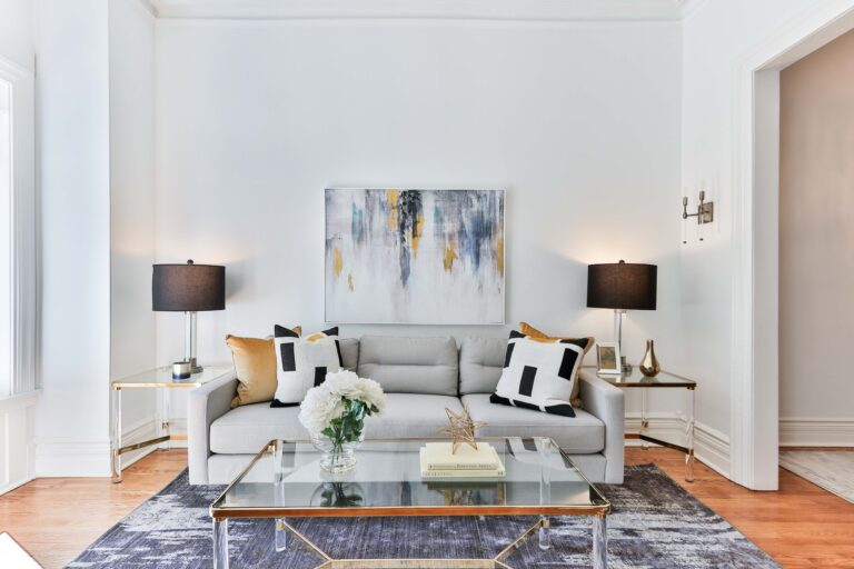 A modern living room perfect for updating your home, featuring a light grey sofa with black and white cushions, abstract wall art, sleek black lamps, a glass coffee table with flowers and gold decor, all atop a dark rug and light wood floors.