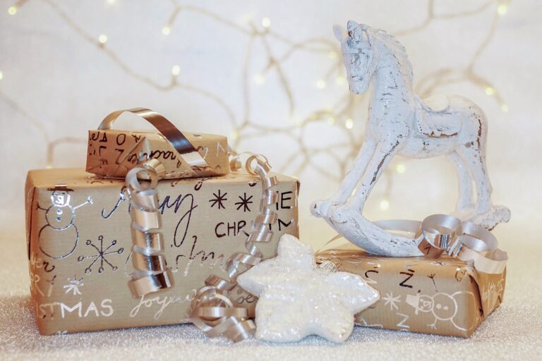 Two gift boxes wrapped in festive paper and silver ribbon sit beside a white decorative rocking horse—perfect Christmas present ideas for children of all ages—with twinkling fairy lights softly glowing in the background.