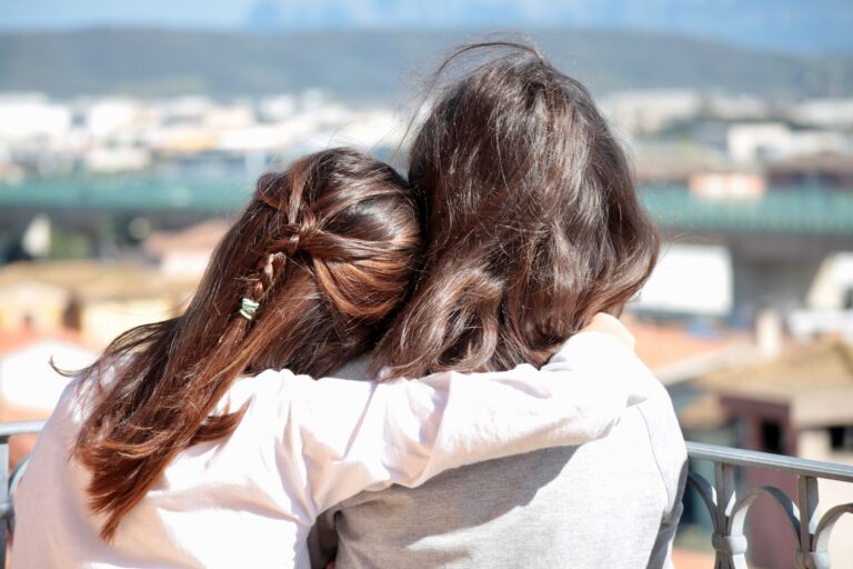 Two people with long brown hair sit side by side, arms round each other, facing away from the camera. As they gaze over a sunny cityscape and distant mountains, they share important life skills to explore with your child from a young age.