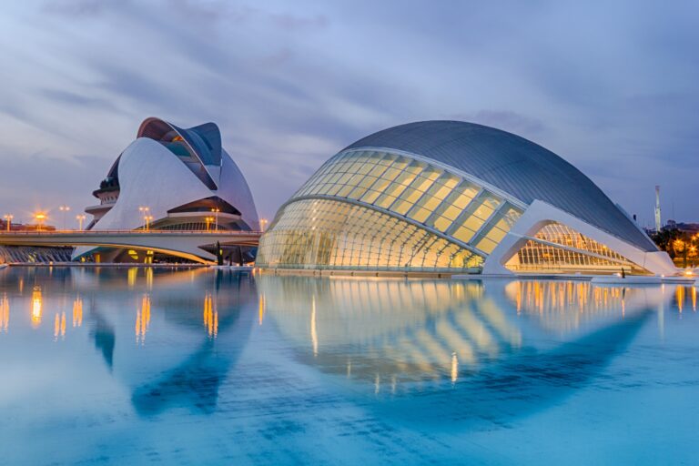 Futuristic buildings with glass and white curves, reflected in a large pool of water at dusk under a cloudy sky—one of the wonderful things to see and taste in Valencia.