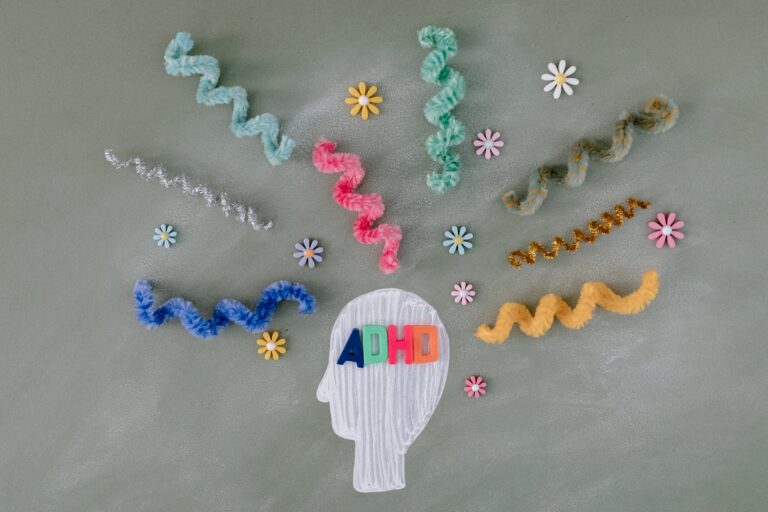 A white outlined head with colourful "ADHD" letters inside is surrounded by pipe cleaners and flower cut-outs on a grey background, creatively illustrating ADHD thoughts or distractions.