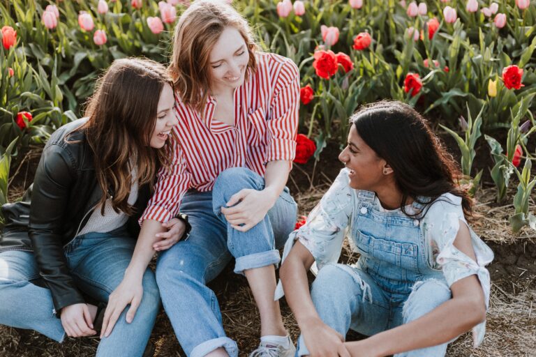Three young women sit on the ground in front of blooming tulips, smiling and laughing together on a sunny day—just like helping your child with their communication skills, they're connecting and enjoying each other’s company in a colourful flower field.