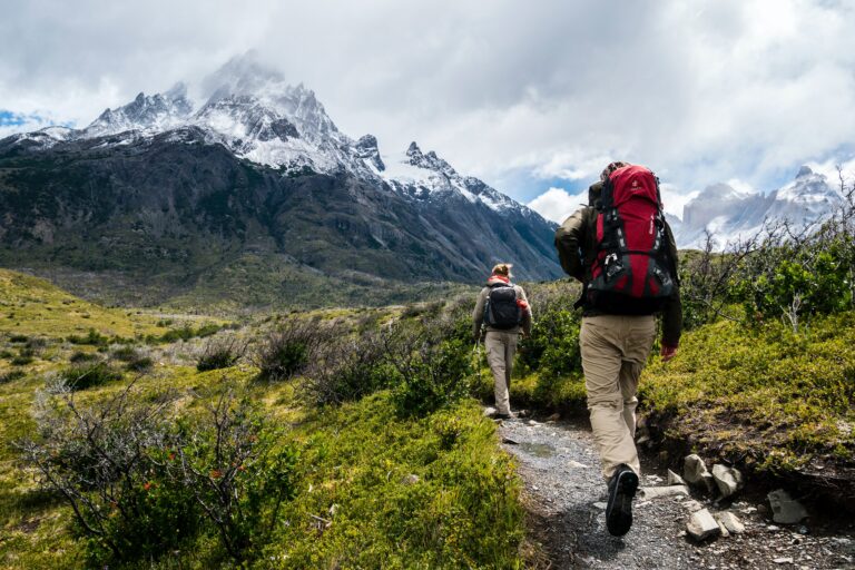 Two hikers with rucksacks walk on a narrow path through green grass and shrubs towards snow-capped mountains under a cloudy sky, illustrating one of the seven reasons why a walking holiday is the perfect holiday to unwind.