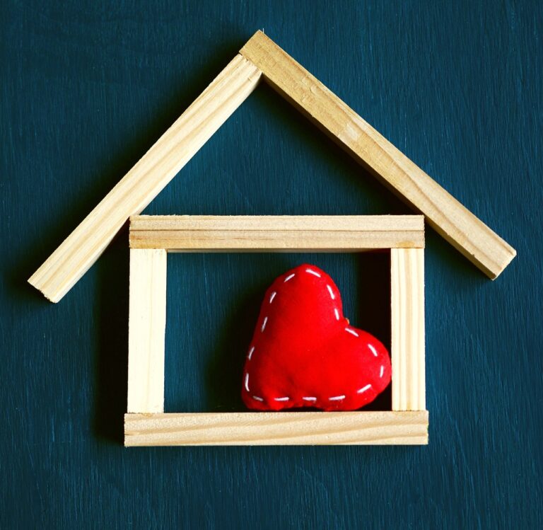 Wooden sticks form the outline of a house with a red fabric heart inside, symbolising love and home, on a dark textured background—inviting you to consider why now is the perfect time to sell your home.