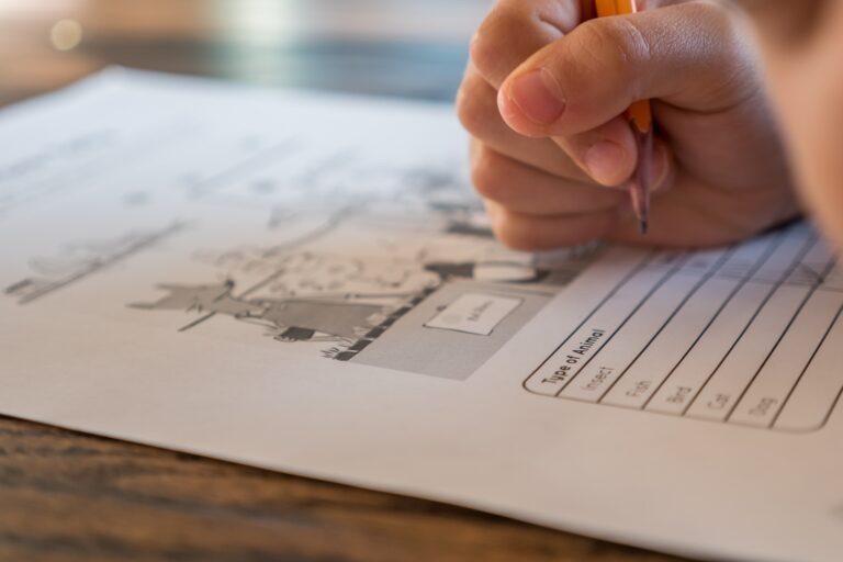 A close-up of a hand holding a pencil, writing on a "Type of Animal" worksheet with printed illustrations and a table, on a wooden surface—an engaging approach for parents learning how to motivate your child to do their homework.
