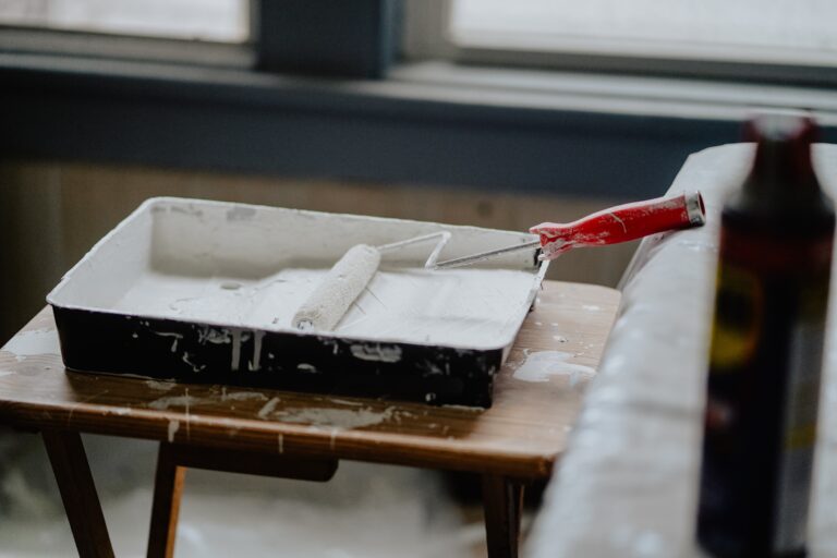 A paint roller with a red handle rests in a tray filled with white paint on a small wooden table—perfect inspiration for your list of 5 Home Improvements to Make in the New Year—with a blurred tin in the foreground and windows in the background.