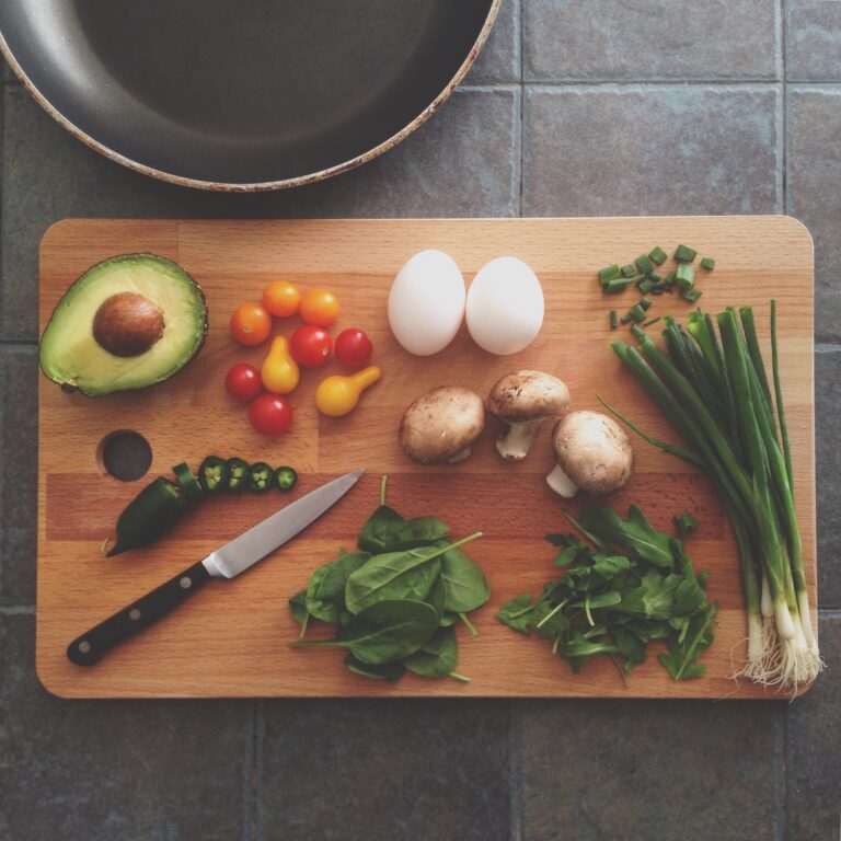 A wooden chopping board with an avocado half, cherry tomatoes, eggs, mushrooms, greens, and a small knife sits on a tiled surface next to a frying pan—showing the fresh ingredients often found when exploring how meal kits work.