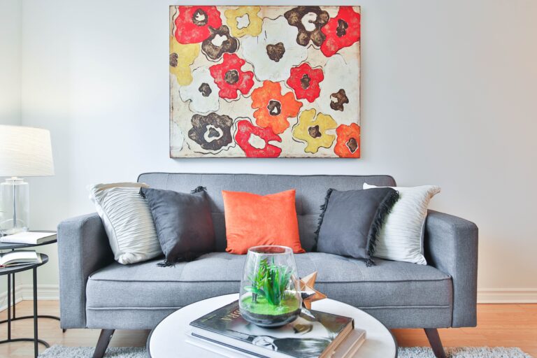 A modern living room offers tips on how to make your living room look bigger and brighter, featuring a grey sofa with four cushions, a round white coffee table with books and a terrarium, and a colourful floral painting above the sofa.