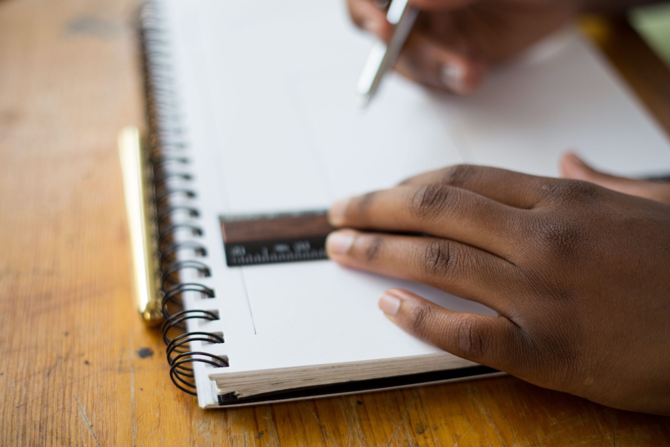 A person uses a pen to draw on a spiral-bound sketchbook, holding a ruler for precision—showcasing the focus needed when setting up the perfect study space for your child. The sketchbook rests on a wooden surface.