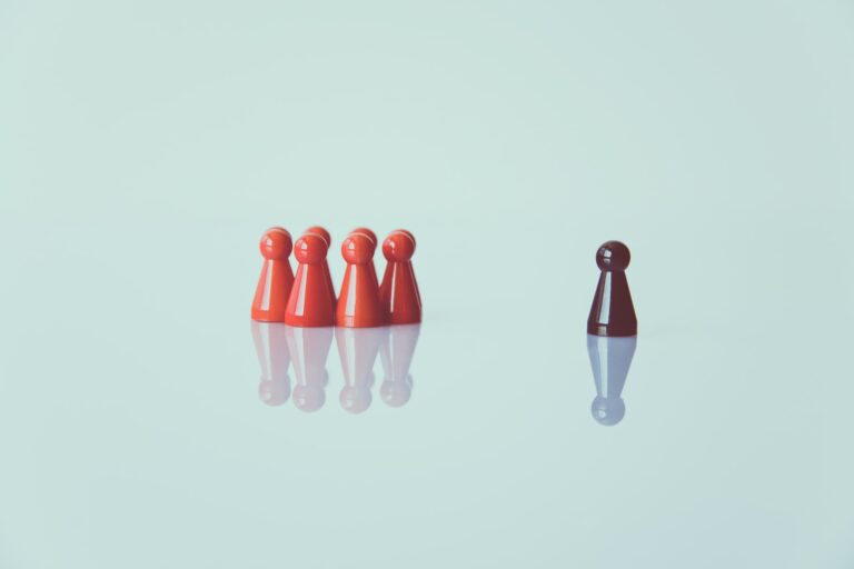 Six red game pieces stand together on a glossy surface, facing a single separated dark red piece on the right—a visual metaphor for teaching your child to resist peer pressure. The background is light and minimalistic.