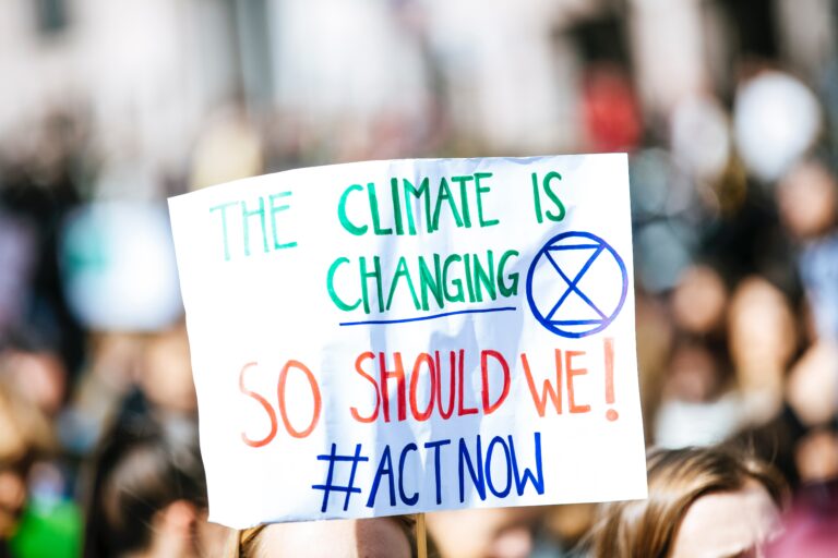 A protester holds a handmade sign reading "The climate is changing, so should we! #ACTNOW" with a blue extinction symbol, amidst a blurred crowd highlighting Three Urgent Climate Actions to Prioritise in 2022.