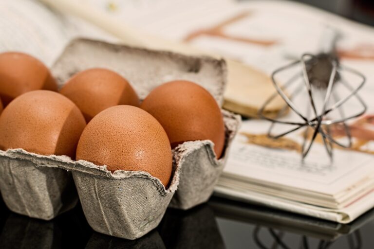A carton of brown eggs sits on a worktop next to an open cookbook featuring 7 Classic Dishes That Can Be Made Gluten-Free, with a metal whisk nearby, suggesting a delicious baking or cooking scene.