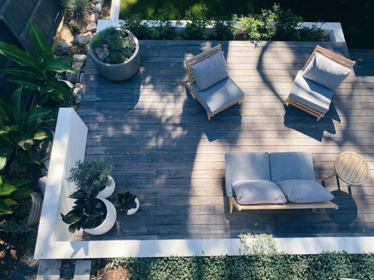 Aerial view of a wooden patio deck, perfect for updating your patio, featuring two single lounge chairs, a double lounge chair, a round side table, and several potted plants, all surrounded by lush greenery and partial shade.