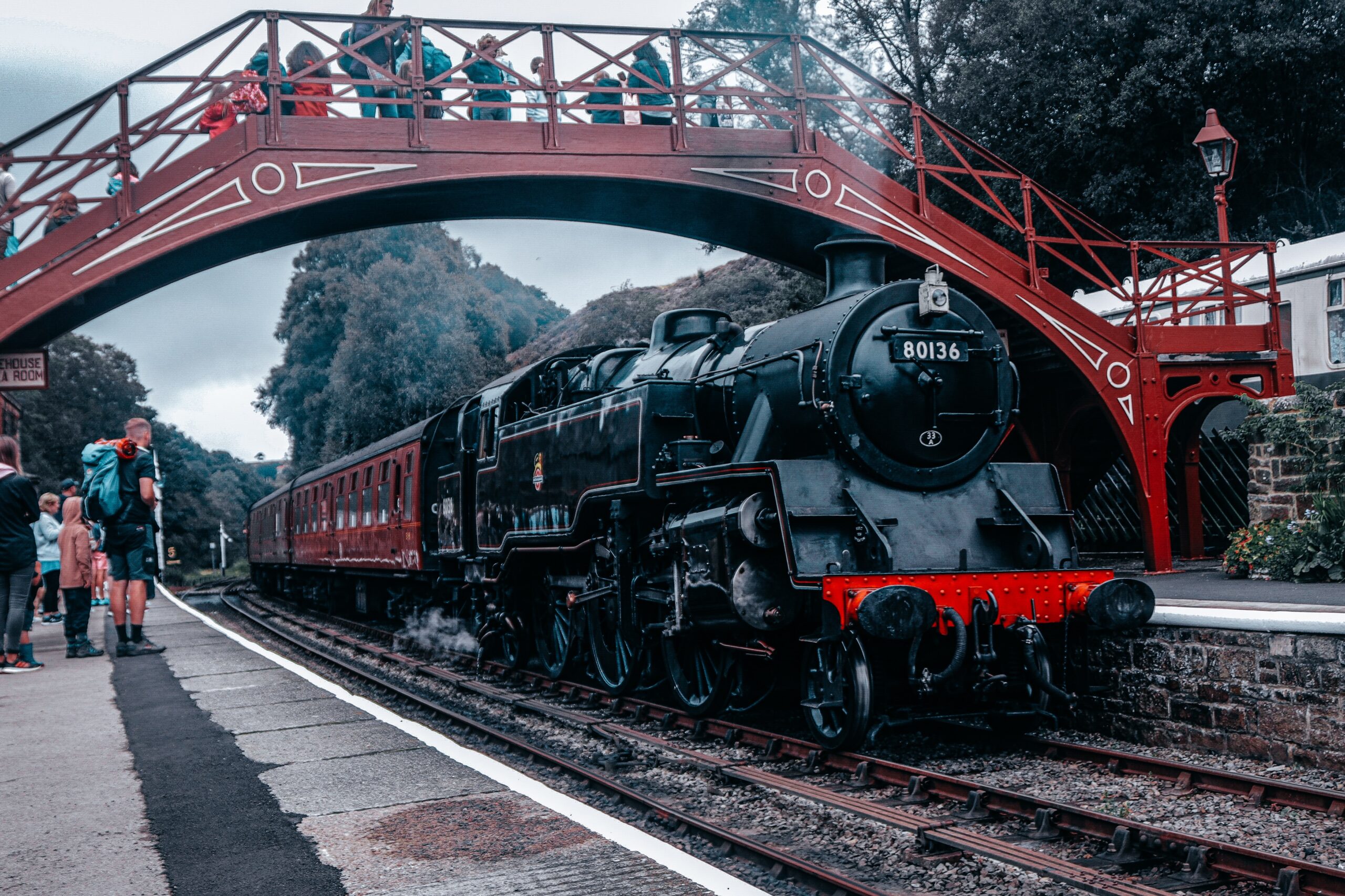 A black steam train with red carriages waits at a station beneath a red footbridge, as people stand on the platform and walk across, surrounded by green trees and overcast skies—one of the Best Train Journeys in the UK.