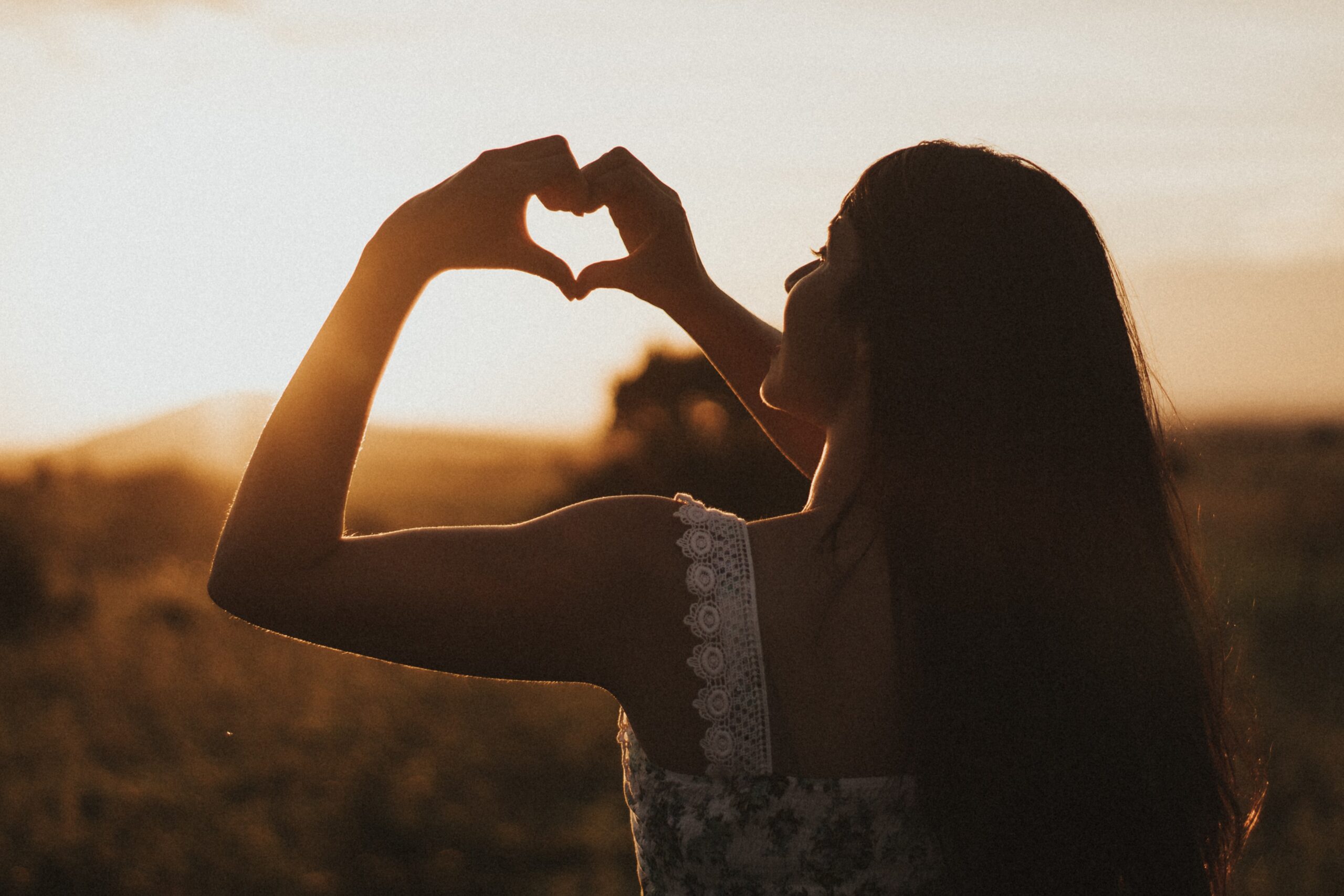 A woman with long hair stands outdoors at sunset, forming a heart shape with her hands against the glowing sky—a moment that inspires self-love and reflects on how to set realistic body goals.