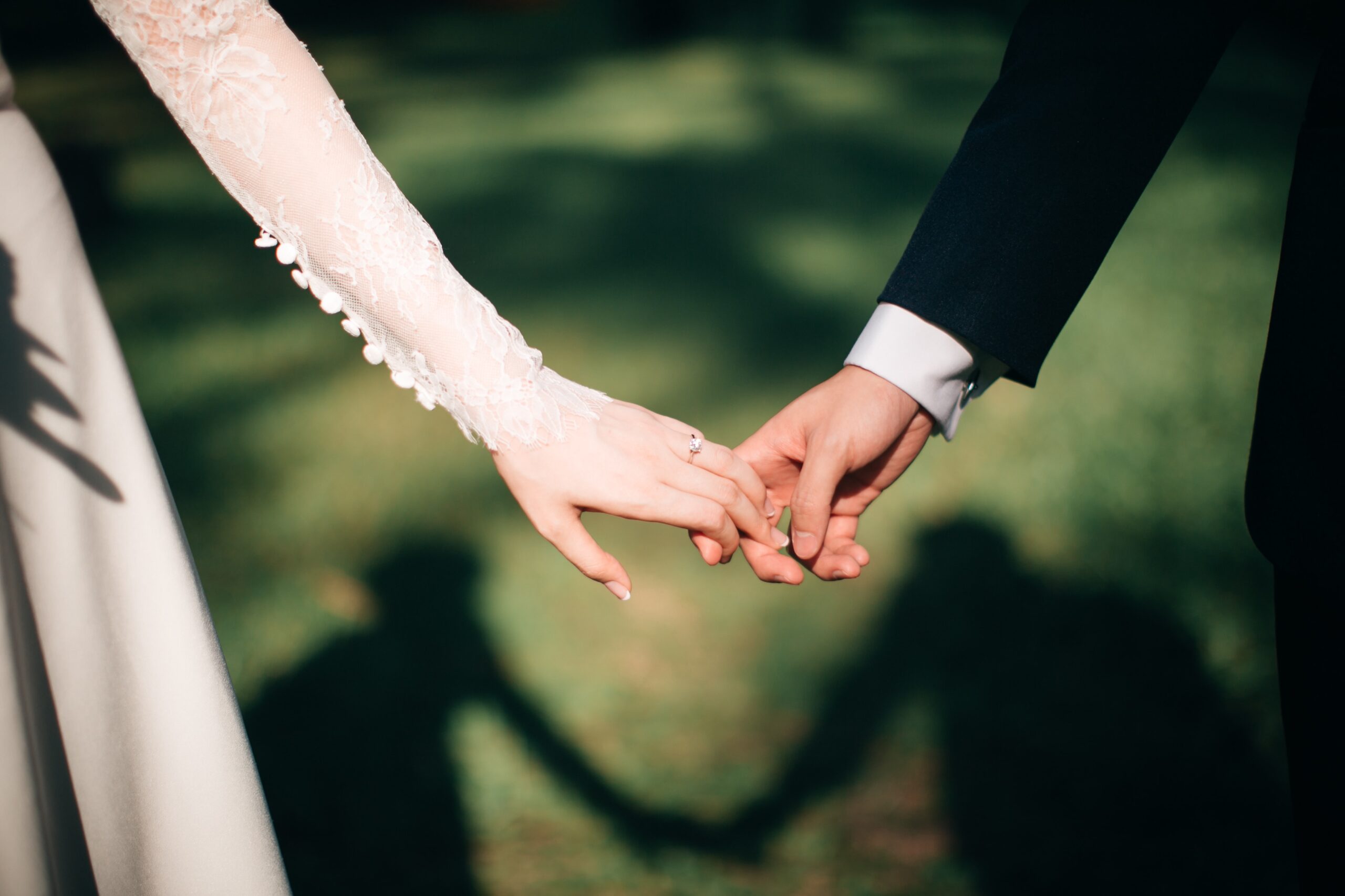 A bride and groom hold hands outdoors, their arms reaching towards each other—a scene straight from Things To Consider When Planning A Wedding In Corsica. The bride’s lace dress and groom’s dark suit cast a heart-shaped shadow on the grass.