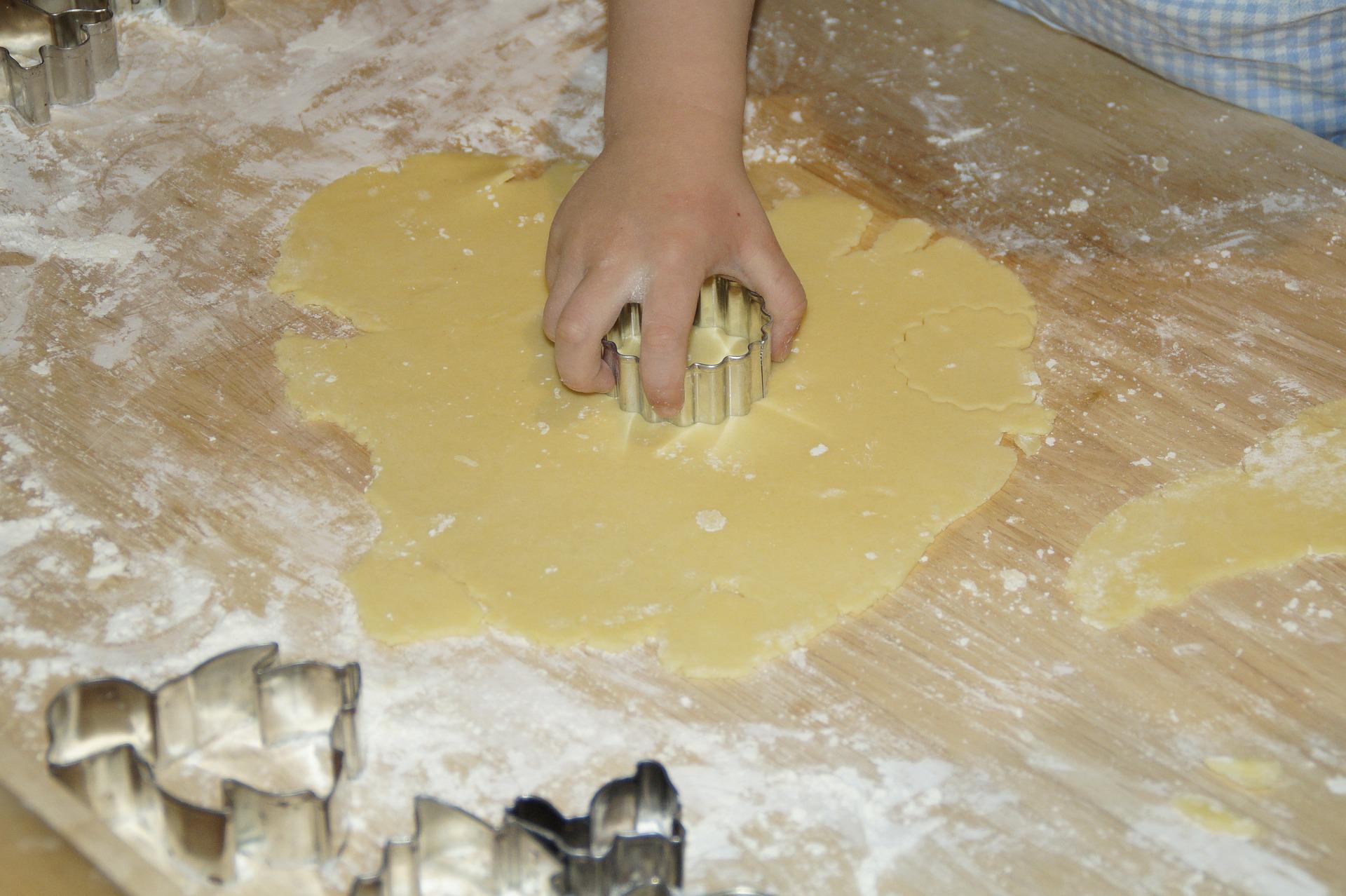 A child’s hand presses a metal biscuit cutter into rolled-out dough on a floured wooden surface, showing how you can involve your children in the kitchen at home. Additional biscuit cutters wait nearby for more fun shapes.