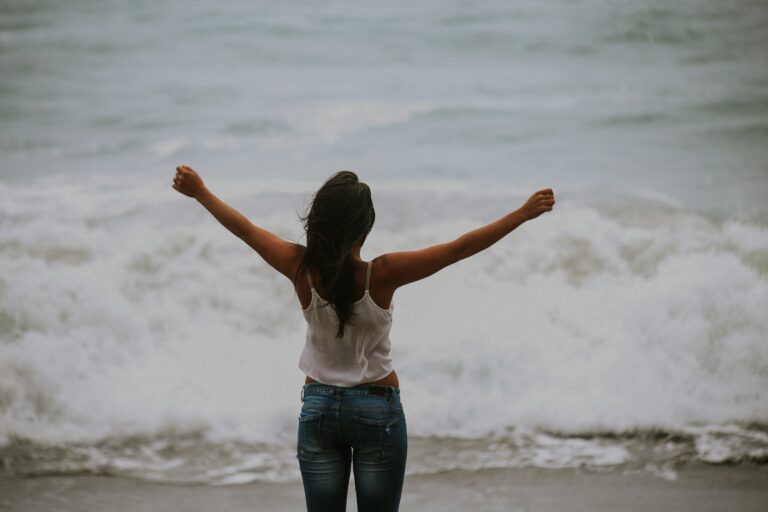 A woman with long hair, wearing a white vest and jeans, stands with arms raised facing the ocean waves, embodying freedom or joy—just like discovering 3 Ways To Deal With Social Anxiety As An Adult.