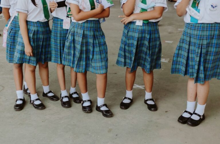 A group of schoolgirls in blue checked skirts, white shirts, and black shoes stand together on a concrete floor, illustrating the benefits of school uniform for children by fostering unity and a sense of belonging.