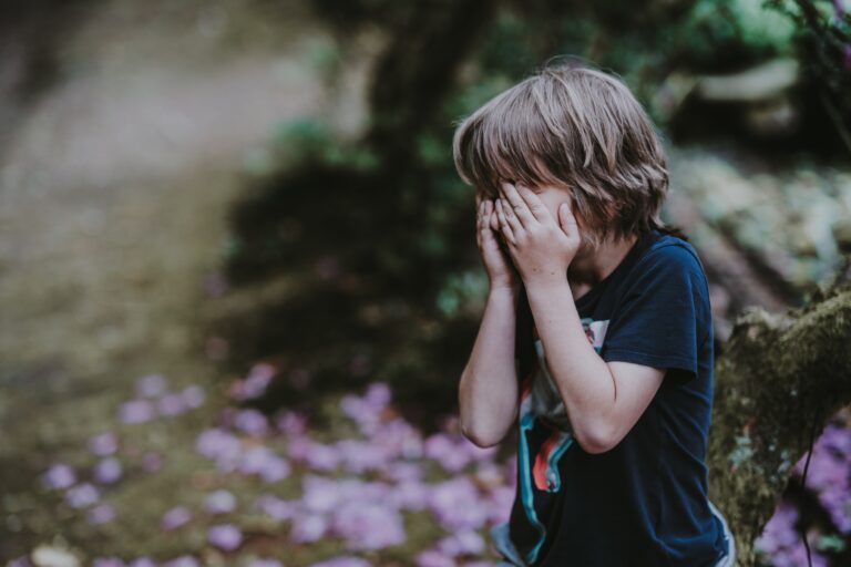 A young child with shoulder-length brown hair covers their face with both hands whilst standing outdoors on a path, surrounded by greenery and scattered pink petals—an authentic moment perfect for teaching your child to manage their emotions.