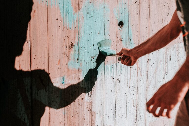 A person’s hand holds a paintbrush, painting light blue strokes on a weathered wooden wall—one of the Simple Home Improvements to Make This Summer—whilst the person’s shadow is also visible on the wall.