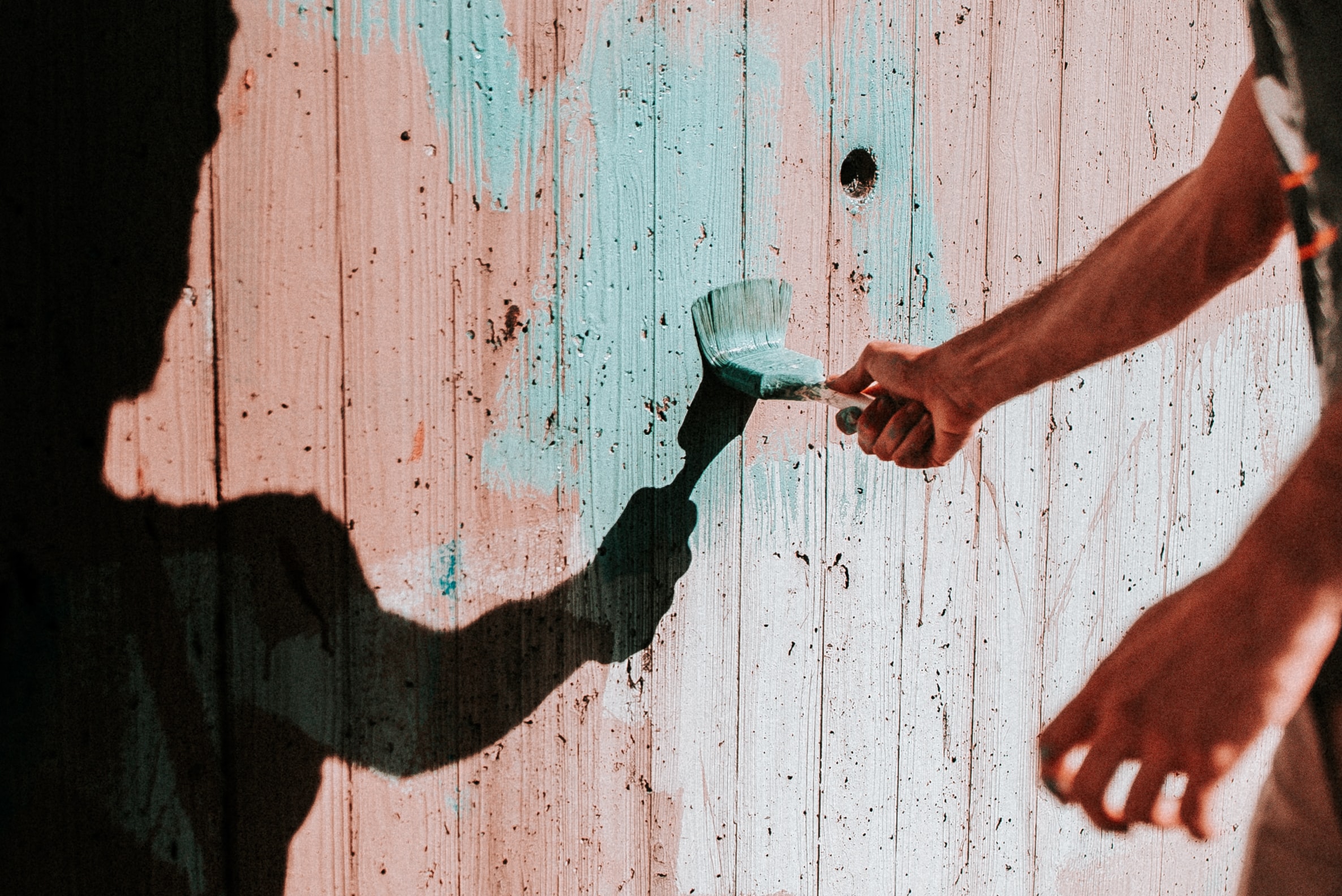 A person’s hand holds a paintbrush, painting light blue strokes on a weathered wooden wall—one of the Simple Home Improvements to Make This Summer—whilst the person’s shadow is also visible on the wall.