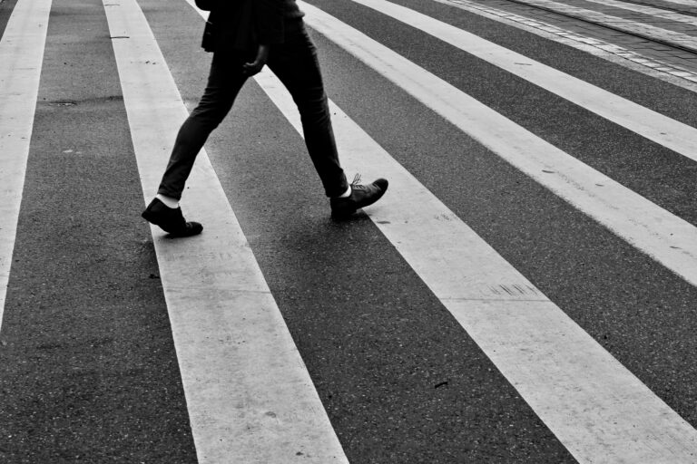 A person wearing dark trousers and shoes walks across a striped zebra crossing. The black and white image, focusing on the legs and feet, subtly highlights the importance of safety—crucial knowledge when learning How To Maximise Your Compensation After A Pedestrian Accident.